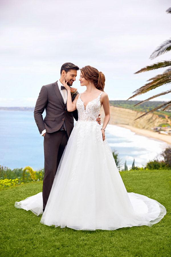 Ein lächelndes Paar im Hochzeitsoutfit steht auf einer grünen Wiese am Meer