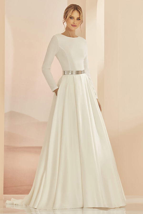 Junge Frau lächelt leicht in elegantem weißen Kleid mit silbernem Gürtel und langen Ärmeln