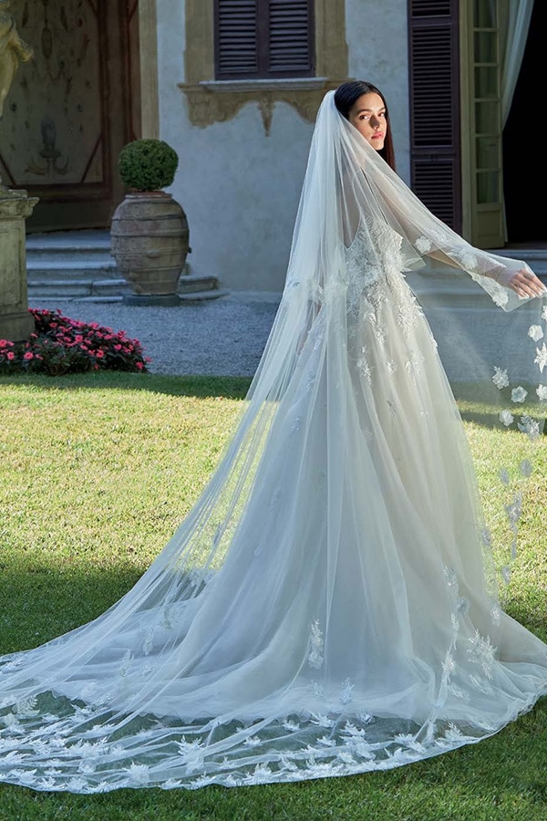 Eine braunhaarige Frau in weißem Spitzenhochzeitskleid mit langem Schleier und ernstem Blick steht im Garten