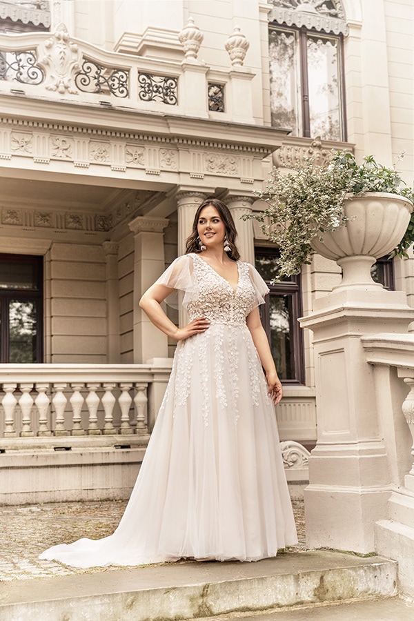 Eine lächelnde Frau im weißen Spitzenkleid steht vor einer eleganten Villa