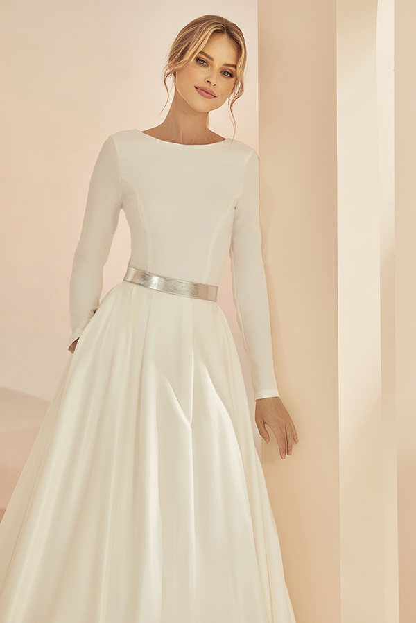 Eine lächelnde Frau mit blonden Haaren trägt ein elegantes weißes Kleid und Silbergürtel