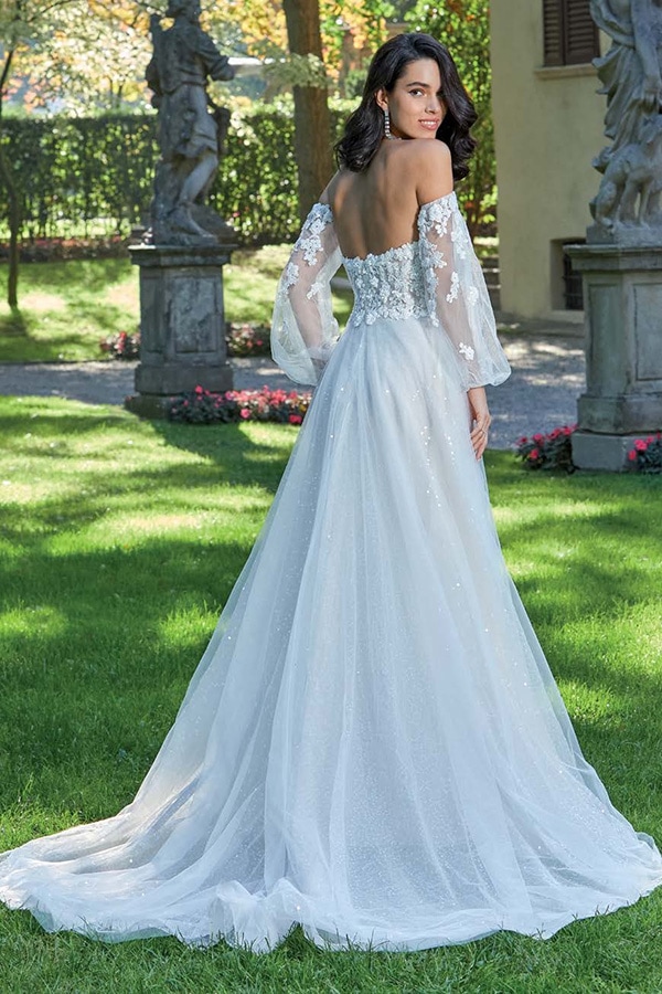 Frau mit dunklem Haar lächelt in einem weißen Brautkleid mit Blumenmuster im Garten