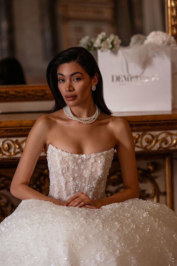 Eine elegante Frau mit langem dunklem Haar in einem glitzernden weißen Brautkleid und Perlenkette sitzt vor einem goldverzierten Spiegel und schaut ruhig