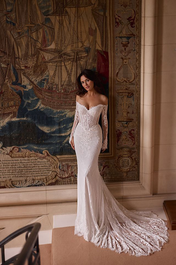 Eine Frau im eleganten weißen Spitzenkleid steht lächelnd vor einem Wandteppich