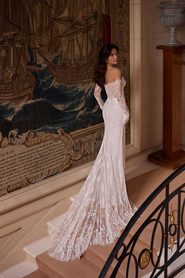 Eine elegant gekleidete Frau im weißen Spitzenkleid mit langen Ärmeln blickt lächelnd zurück