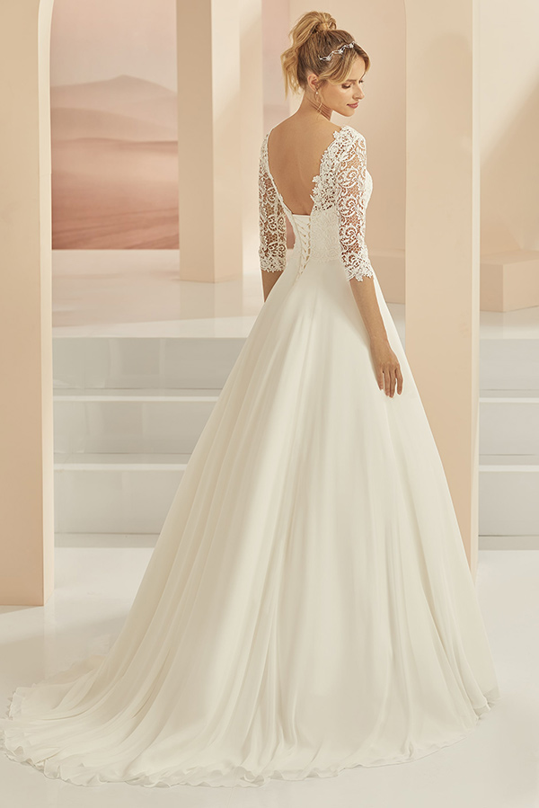 Frau mit Hochsteckfrisur in weißem Spitzenbrautkleid lächelt zart und blickt zurück