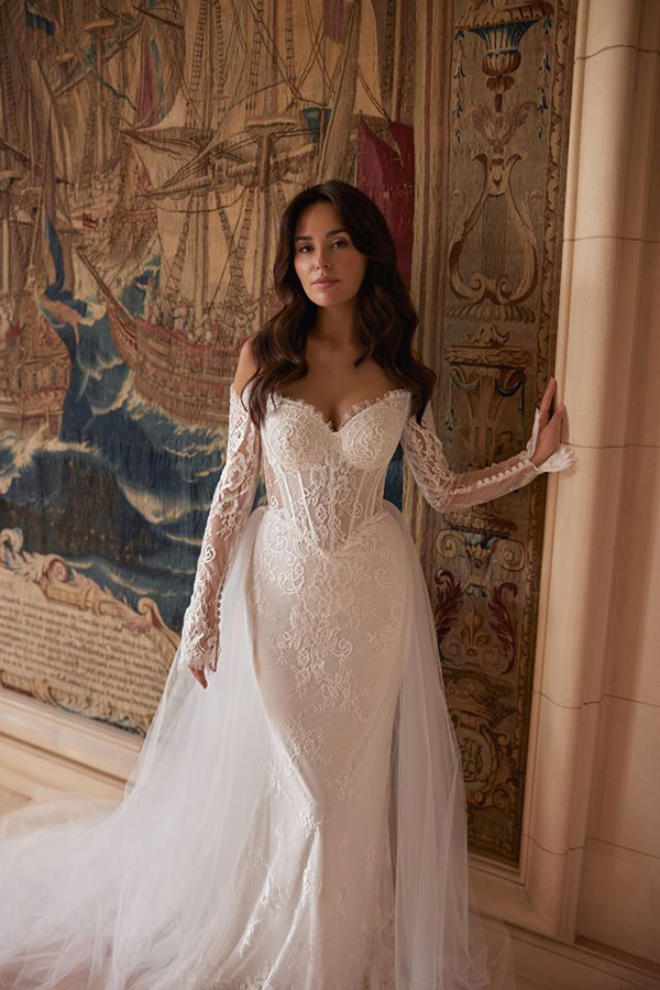 Junge Frau mit langen braunen Haaren in eleganter Spitze Brautkleid steht lächelnd vor einem Wandteppich mit historischen Schiffen und warmen Farbtönen