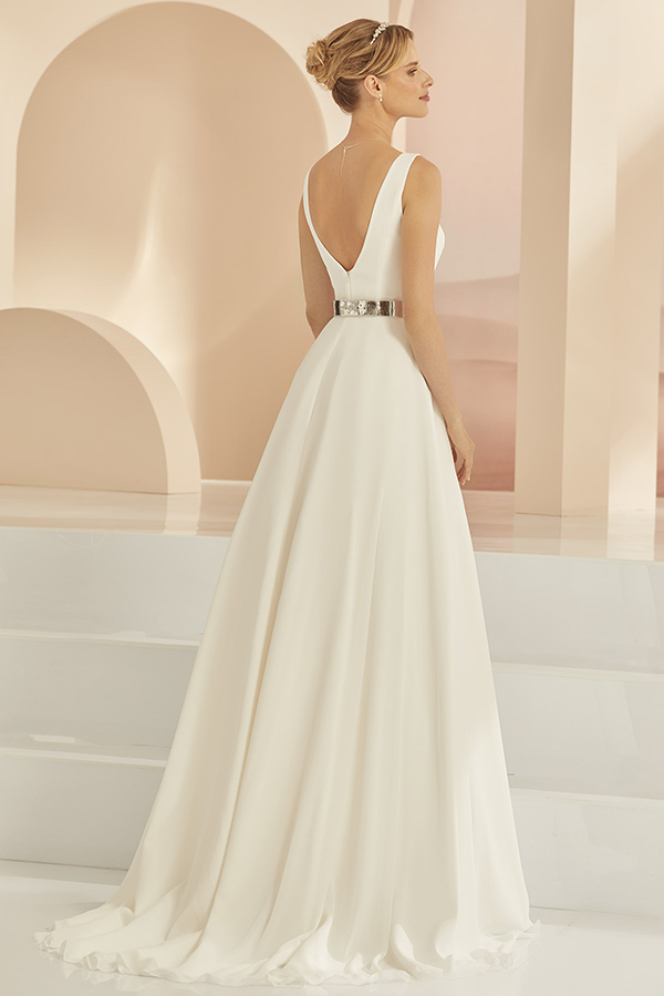 Eine lächelnde Frau im weißen eleganten Abendkleid mit silbernem Gürtel und schlichter Frisur vor pastellfarbenen Bögen