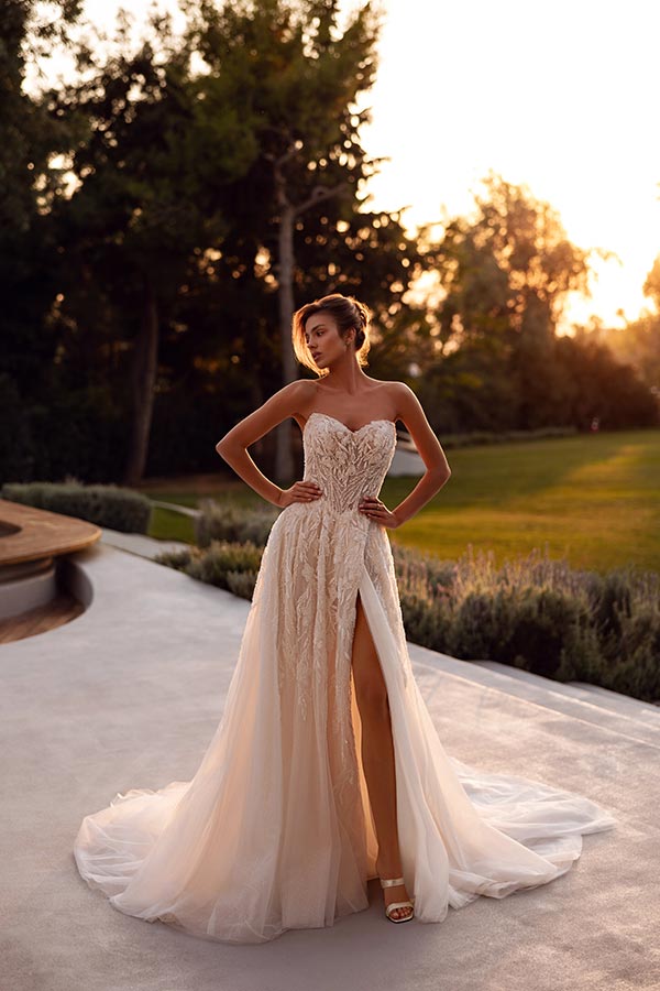 Eine junge Frau im cremefarbenen Brautkleid mit Schlitz steht selbstbewusst im Garten