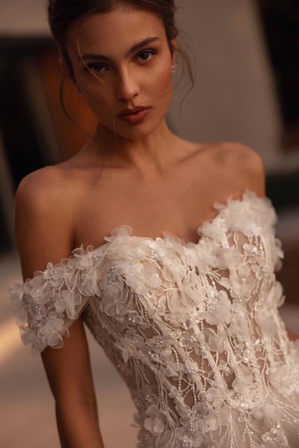 Junge Frau mit ernstem Blick trägt ein weißes, florales Brautkleid mit 3D-Blütenornamenten