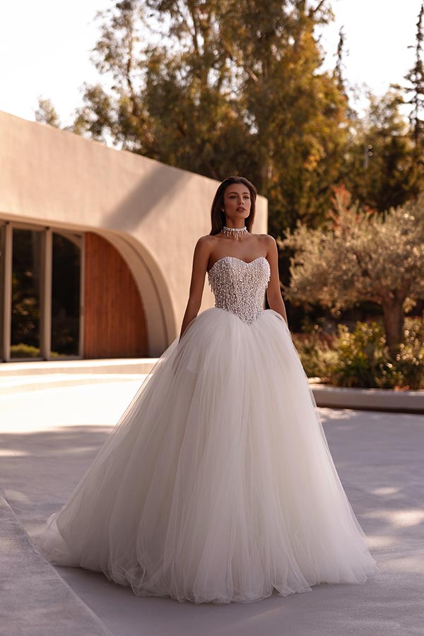 Eine junge Frau mit ernstem Blick trägt ein prachtvolles, weißes Brautkleid draußen