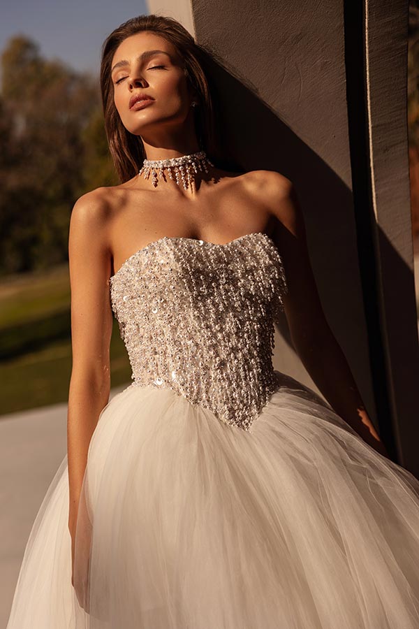 Junge Frau mit funkelndem, trägerlosem Hochzeitskleid und Perlenhalskette steht entspannt im Sonnenlicht