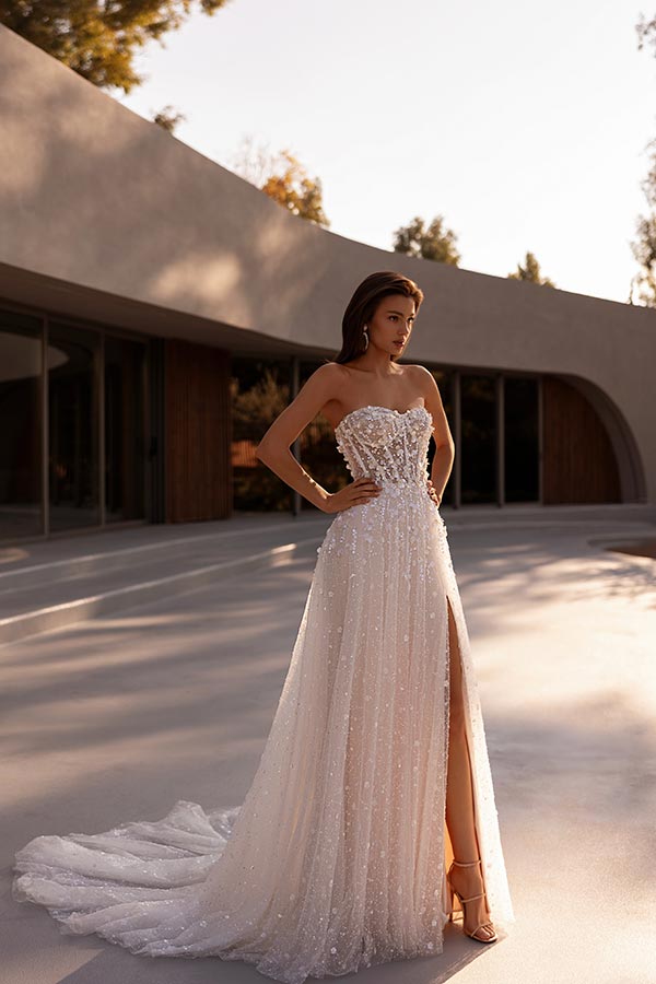 Junge Frau im glitzernden weißen Brautkleid mit Schlitz steht entspannt im Sonnenschein