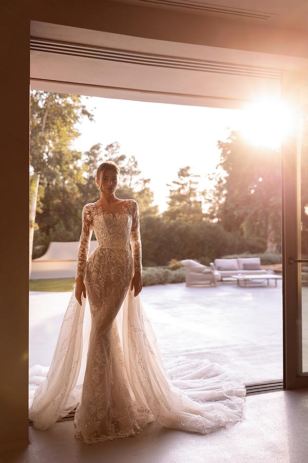 Eine elegant gekleidete Frau im Spitzenkleid mit Schleier steht lächelnd im Sonnenlicht