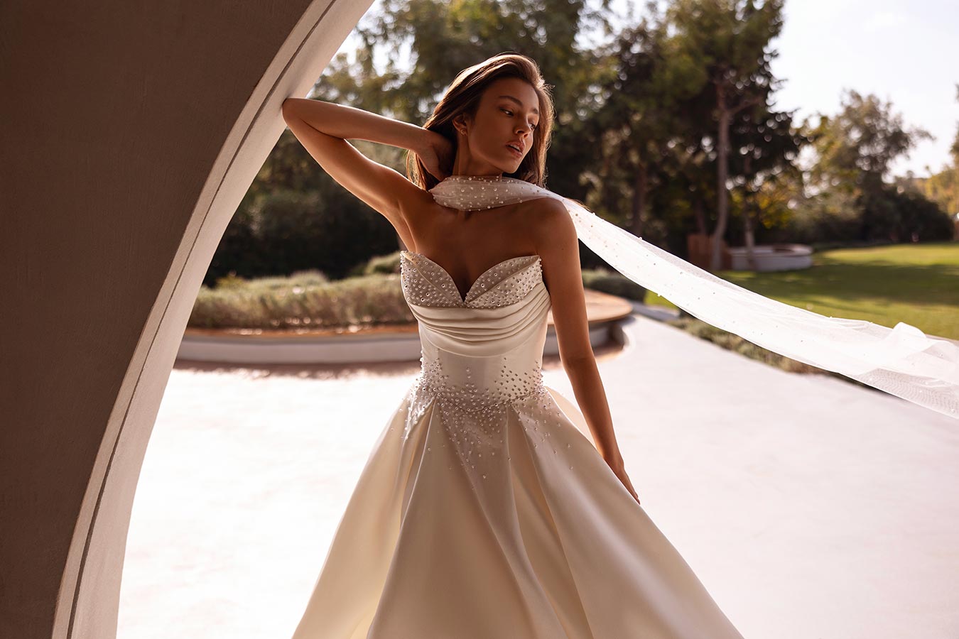 Eine Frau in cremeweissem Brautkleid steht elegant draussen im Sonnenlicht