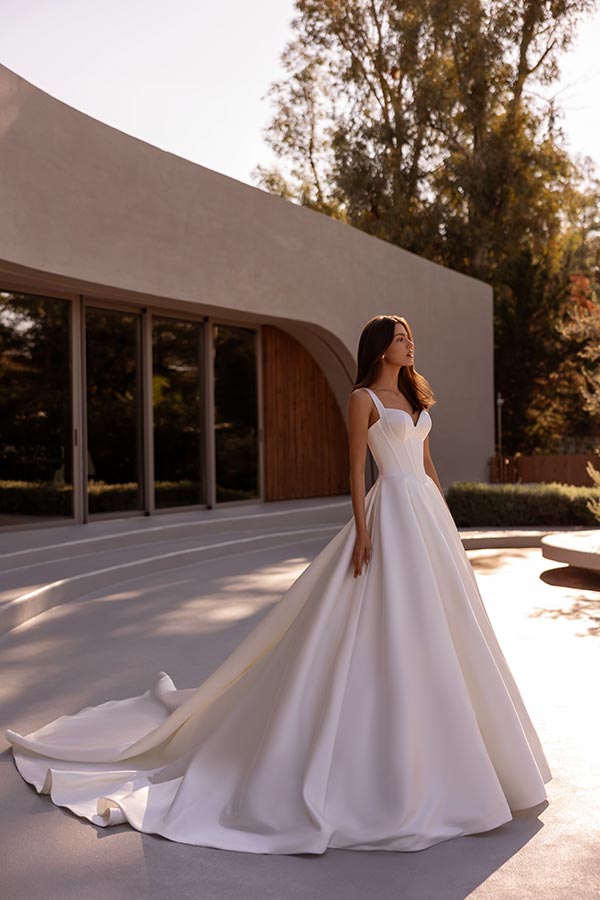 Eine lächelnde Frau im weißen Brautkleid steht vor einem modernen Gebäude im Sonnenschein