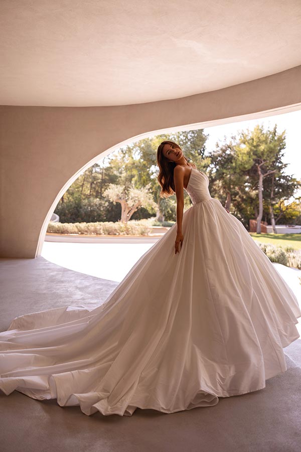 Eine lächelnde Frau im weißen Brautkleid steht mit weitem Rock im sonnigen Raum