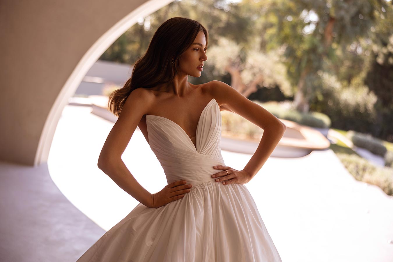 Eine junge Frau mit langen braunen Haaren im eleganten weißen Brautkleid steht draußen