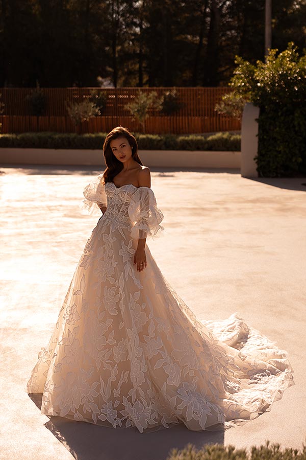 Eine Frau mit langem braunen Haar lächelt leicht und trägt ein elegantes cremefarbenes Spitzenkleid