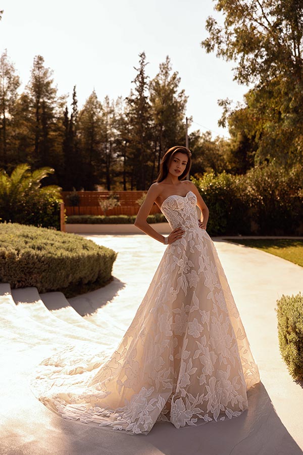 Eine Frau mit ernstem Blick steht im Sonnenlicht in einem eleganten cremefarbenen Kleid mit Spitzenmustern