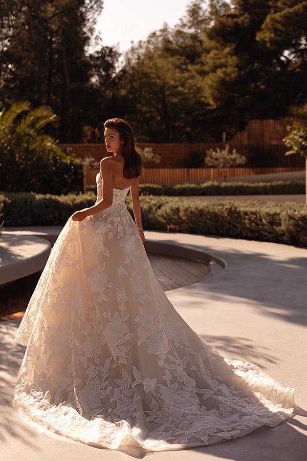 Eine Frau mit offenem Haar lächelt und trägt ein elegantes cremefarbenes Brautkleid mit floralen Mustern im Sonnenlicht