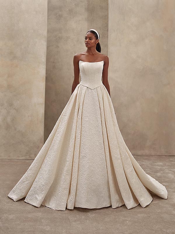 Eine dunkelhaarige Frau in einem cremefarbenen Brautkleid steht entspannt und lächelt leicht