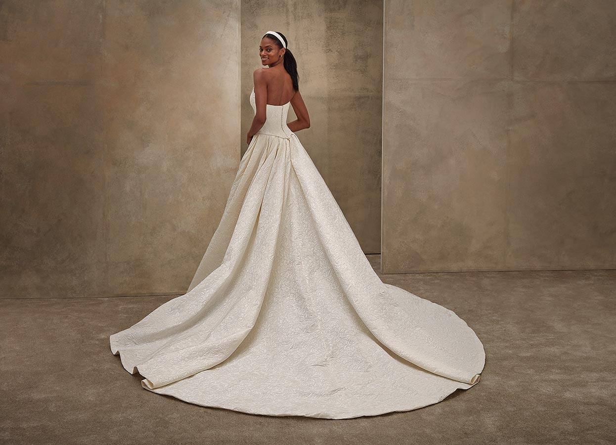 Eine lächelnde Frau mit dunkler Haut im eleganten cremefarbenen Brautkleid mit langer Schleppe und Haarband vor beigem Hintergrund