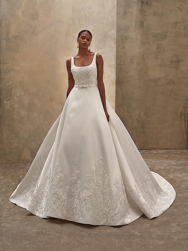 Junge Frau mit ruhigem Gesichtsausdruck in elegantem cremefarbenen Brautkleid mit Spitzenapplikationen und weiter Schleppe vor beigem Hintergrund