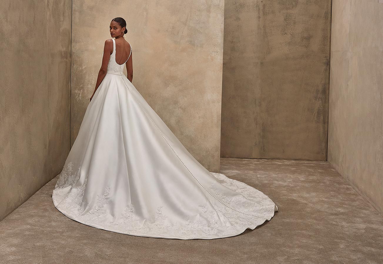 Eine elegant gekleidete Frau in weißem Brautkleid steht lächelnd vor beigem Hintergrund