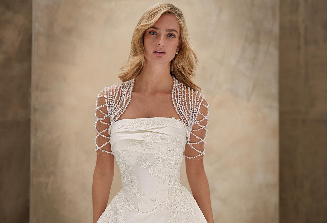 Eine blonde Frau mit sanftem Blick trägt ein weißes Brautkleid mit Perlenbolero