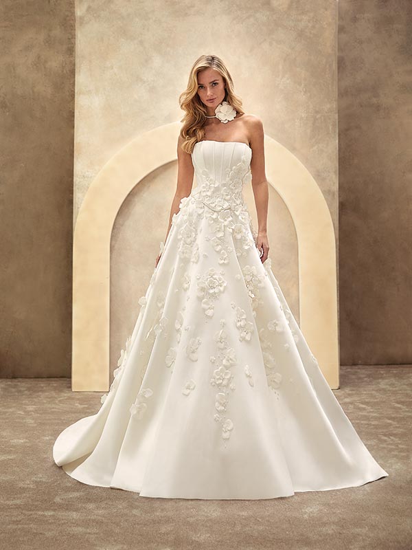 Eine blonde Frau mit ernster Miene trägt ein elegantes, weißes Brautkleid mit 3D-Blumenornamenten vor beiger Kulisse