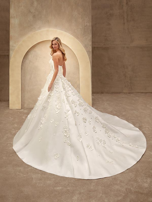 Eine blonde Frau blickt lächelnd über die Schulter, trägt ein weißes Brautkleid mit floralen Applikationen und steht vor einem cremefarbenen Bogen auf sandfarbener Kulisse