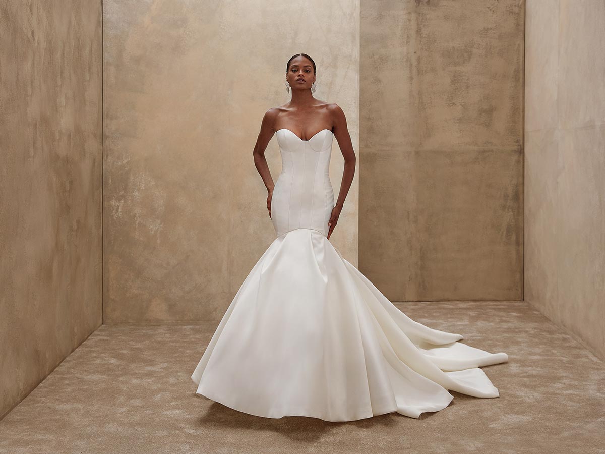 Eine elegante Frau in weißem, schulterfreiem Brautkleid steht selbstbewusst in neutralem Raum