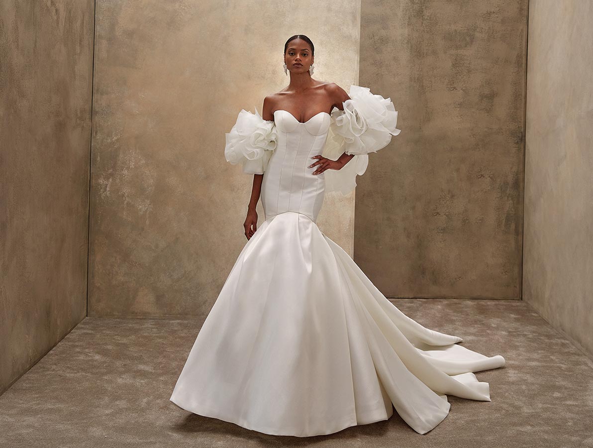 Eine elegante Frau mit ernstem Gesichtsausdruck trägt ein weißes Kleid mit voluminösen Ärmeln