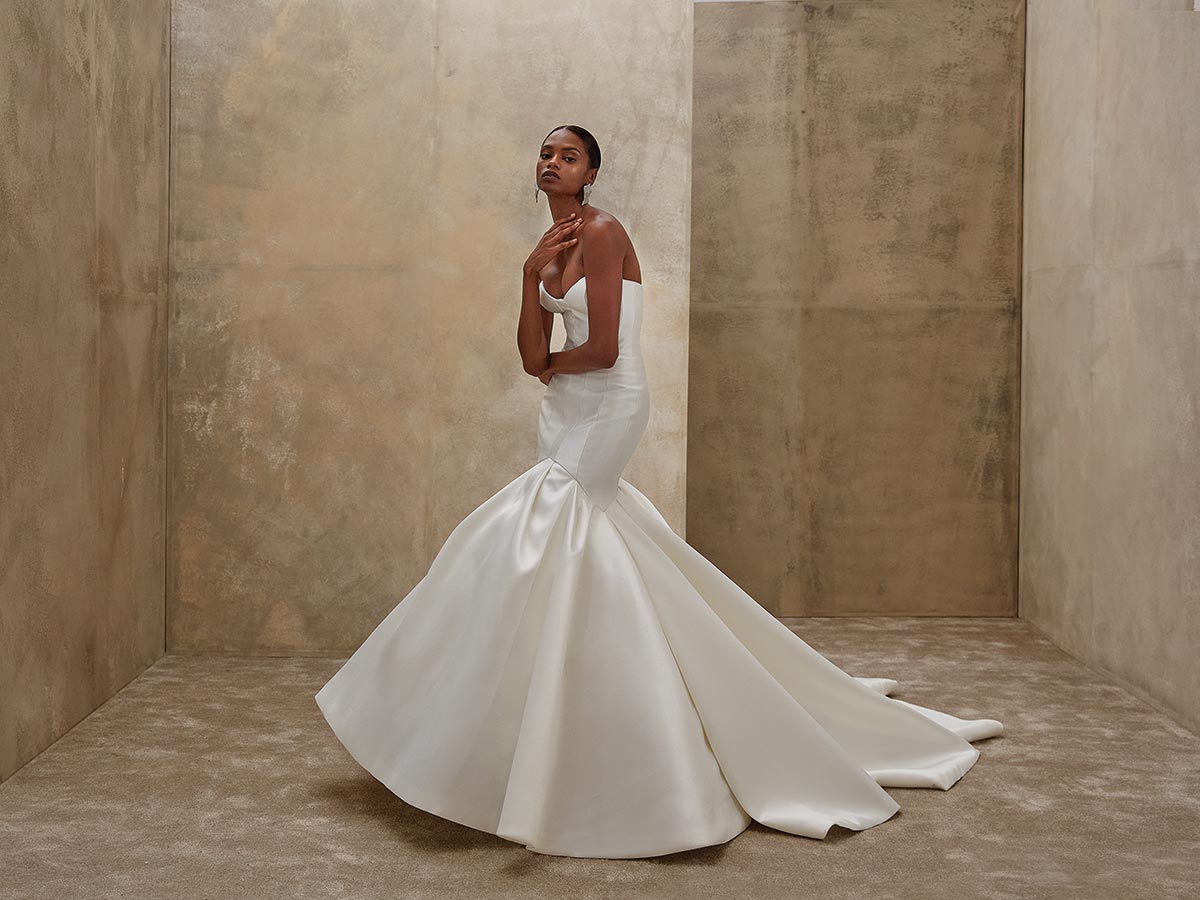 Eine elegante Frau im weißen Brautkleid steht ernst in einem goldbeigen Raum