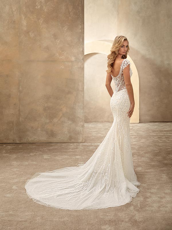 Eine blonde Frau blickt ernst über die Schulter, trägt ein elegantes cremefarbenes Spitzenkleid mit langer Schleppe und durchsichtigen Rückendetails