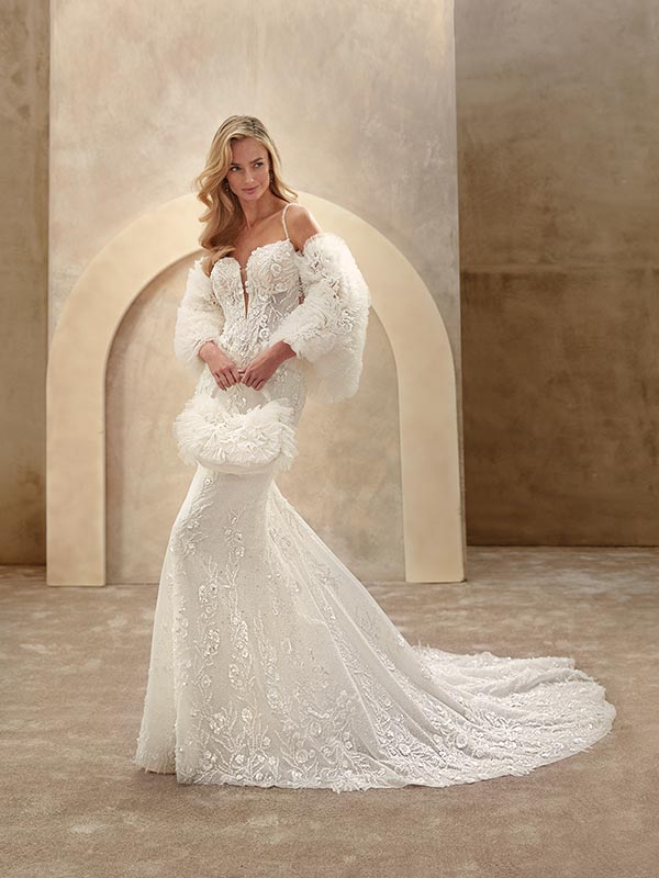 Eine blonde Frau lächelt leicht in einem weißen Spitzenkleid mit langem Schleier und Federn