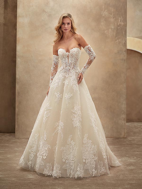 Eine blonde Frau mit ernstem Blick trägt ein cremefarbenes Brautkleid mit Spitze