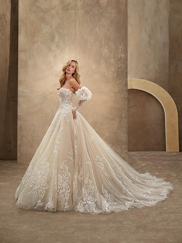 Eine blonde Frau lächelt sanft in einem elfenbeinfarbenen Spitzenbrautkleid mit langen Ärmeln