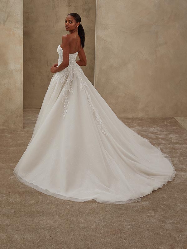 Eine Frau mit langem dunklem Haar trägt ein weißes Brautkleid mit Spitzenapplikationen und schaut mit ernstem Blick über die Schulter