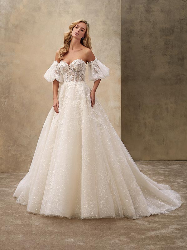 Eine blonde Frau in einem eleganten cremefarbenen Brautkleid mit Spitzenärmeln und schimmernden Details steht entspannt vor einem neutralen Hintergrund