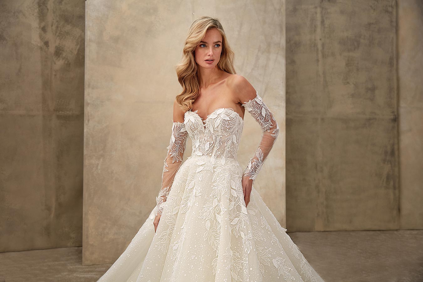 Eine blonde Frau mit welligem Haar trägt ein elegantes cremefarbenes Brautkleid