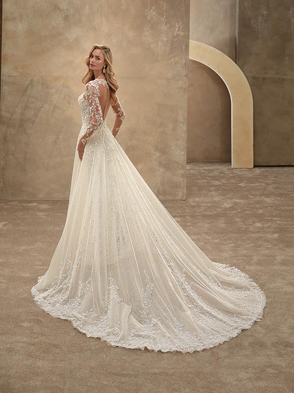 Eine blonde Frau mit langem Haar trägt ein elegantes Spitzenkleid mit langer Schleppe und lächelt sanft