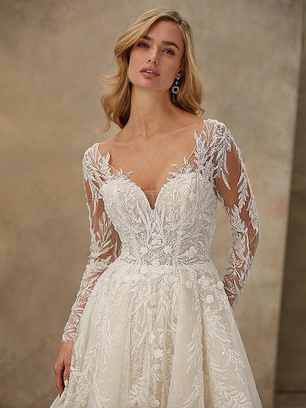 Eine blonde Frau lächelt sanft in einem hellen Spitzenhochzeitskleid mit langen Ärmeln