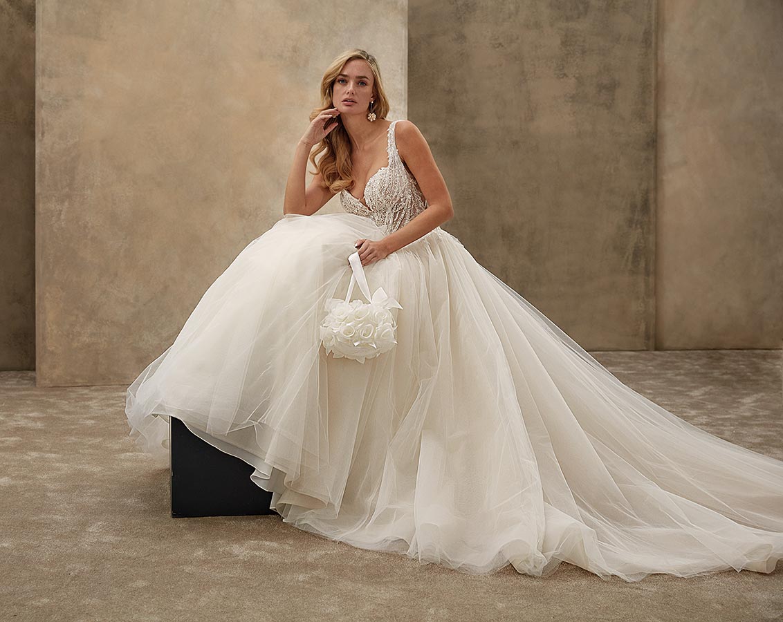 Eine blonde Frau im weißen Brautkleid sitzt nachdenklich mit Blumenkorb auf Kasten