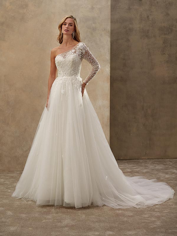 Eine blonde Frau steht ruhig in einem weißen, einarmigen Brautkleid mit Spitze