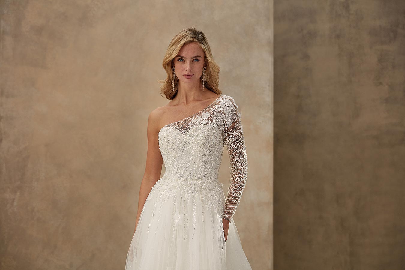 Eine blonde Frau lächelt sanft in einem eleganten Ein-Schulter-Brautkleid mit Spitze