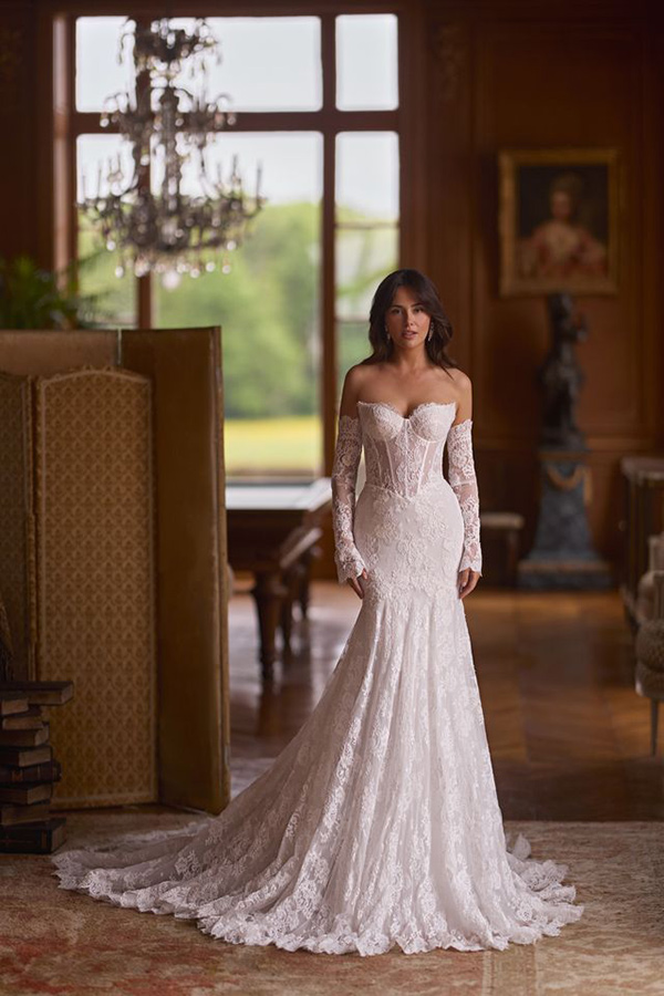 Eine Frau mit ernstem Ausdruck trägt ein elegantes weißes Spitzenkleid mit langen Ärmeln und steht in einem prunkvollen Raum mit großem Fenster