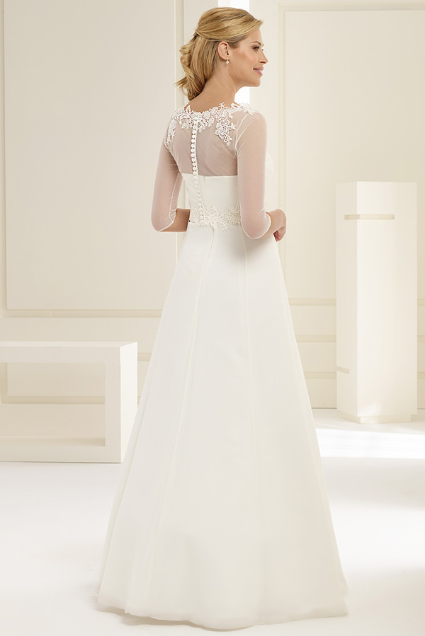 Eine blonde Frau im weißen Hochzeitskleid mit Spitzenärmeln steht lächelnd im hellen Raum