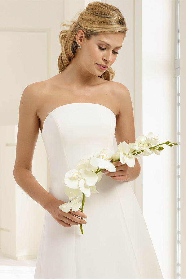 Eine blonde Frau im weißen Brautkleid hält weiße Orchideen und blickt sanft nach unten